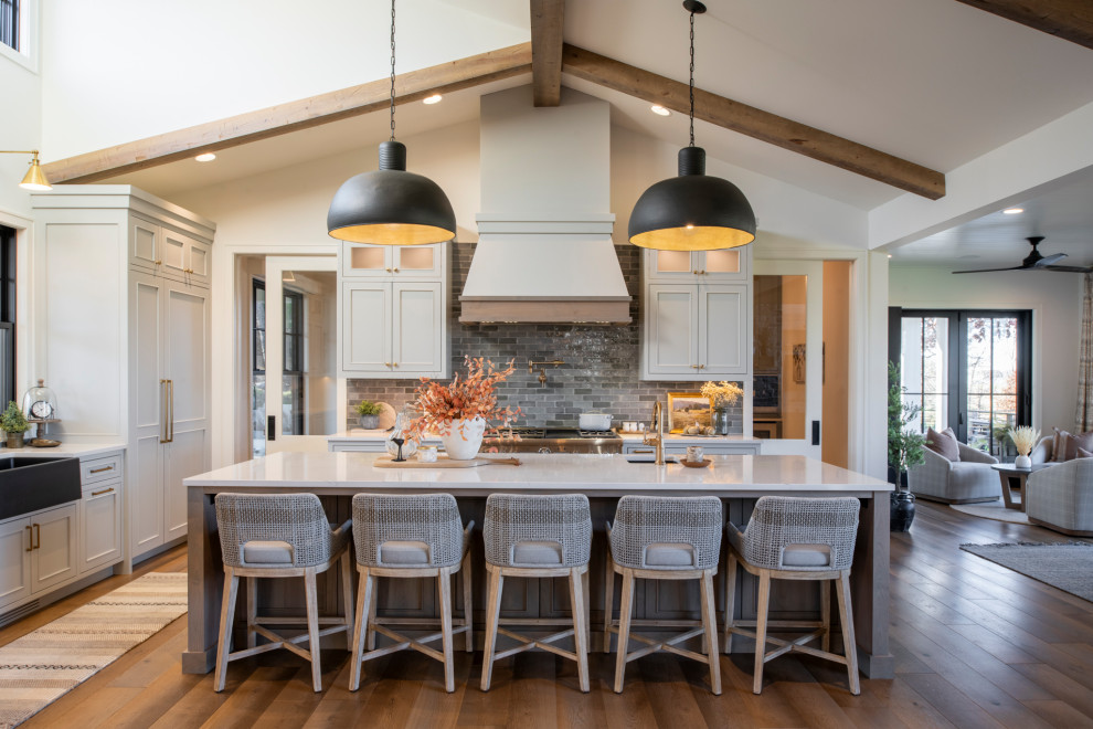 Example of a transitional l-shaped dark wood floor, brown floor, exposed beam and vaulted ceiling kitchen design in Minneapolis with a farmhouse sink, recessed-panel cabinets, white cabinets, gray backsplash, stainless steel appliances, an island and white countertops