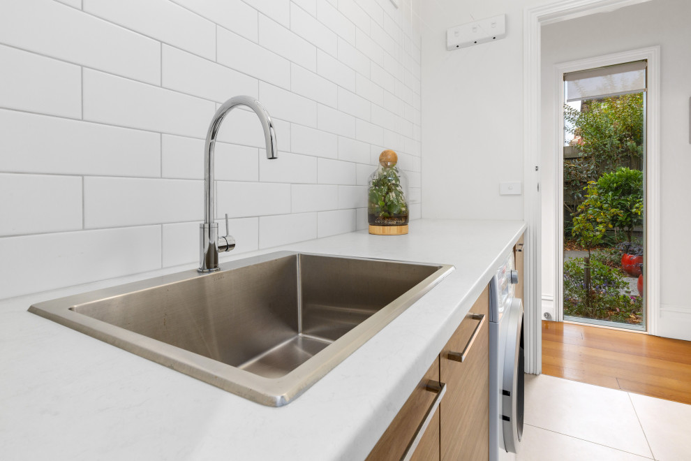 This is an example of a mid-sized scandinavian galley dedicated laundry room in Melbourne with an utility sink, flat-panel cabinets, light wood cabinets, quartz benchtops, white splashback, subway tile splashback, white walls, porcelain floors, a concealed washer and dryer, beige floor and white benchtop.