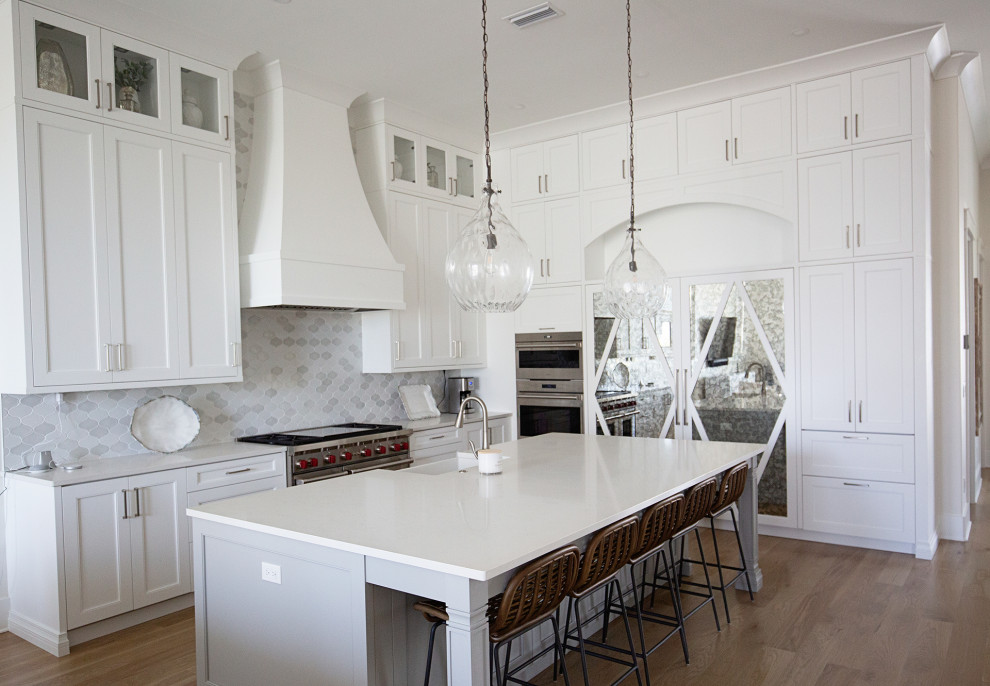 Photo of a large transitional l-shaped eat-in kitchen in Miami with a farmhouse sink, shaker cabinets, white cabinets, quartzite benchtops, white splashback, ceramic splashback, stainless steel appliances, medium hardwood floors, with island, beige floor, white benchtop and recessed.