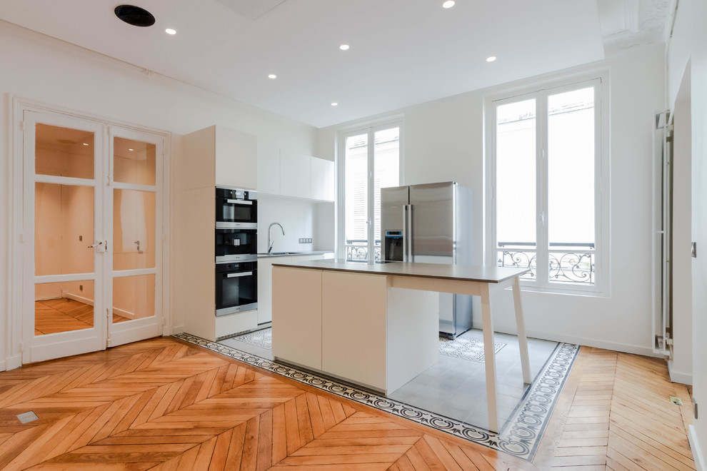 This is an example of a mid-sized modern single-wall open plan kitchen in Paris with an integrated sink, flat-panel cabinets, white cabinets, granite benchtops, white splashback, glass sheet splashback, stainless steel appliances, cement tiles, with island, grey floor and grey benchtop.