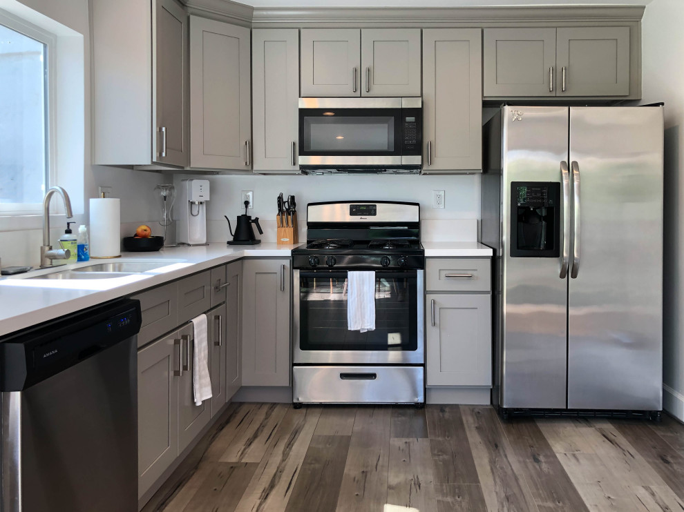 Photo of a mid-sized scandinavian l-shaped eat-in kitchen in Los Angeles with shaker cabinets, grey cabinets, stainless steel appliances, no island, brown floor, white benchtop, granite benchtops, white splashback, granite splashback, laminate floors and a double-bowl sink.