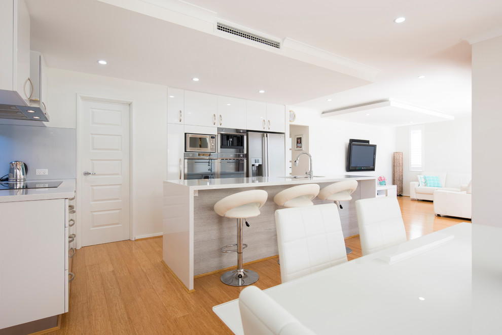 This is an example of a mid-sized contemporary l-shaped open plan kitchen in Perth with an undermount sink, white cabinets, solid surface benchtops, white splashback, glass sheet splashback, stainless steel appliances, bamboo floors and with island.