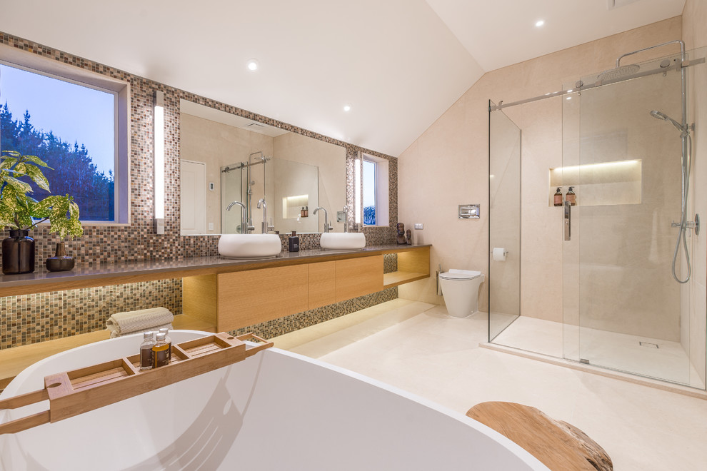Large contemporary master bathroom in Auckland with flat-panel cabinets, light wood cabinets, a freestanding tub, a corner shower, a one-piece toilet, beige tile, limestone, beige walls, limestone floors, a vessel sink, engineered quartz benchtops, beige floor and a sliding shower screen.