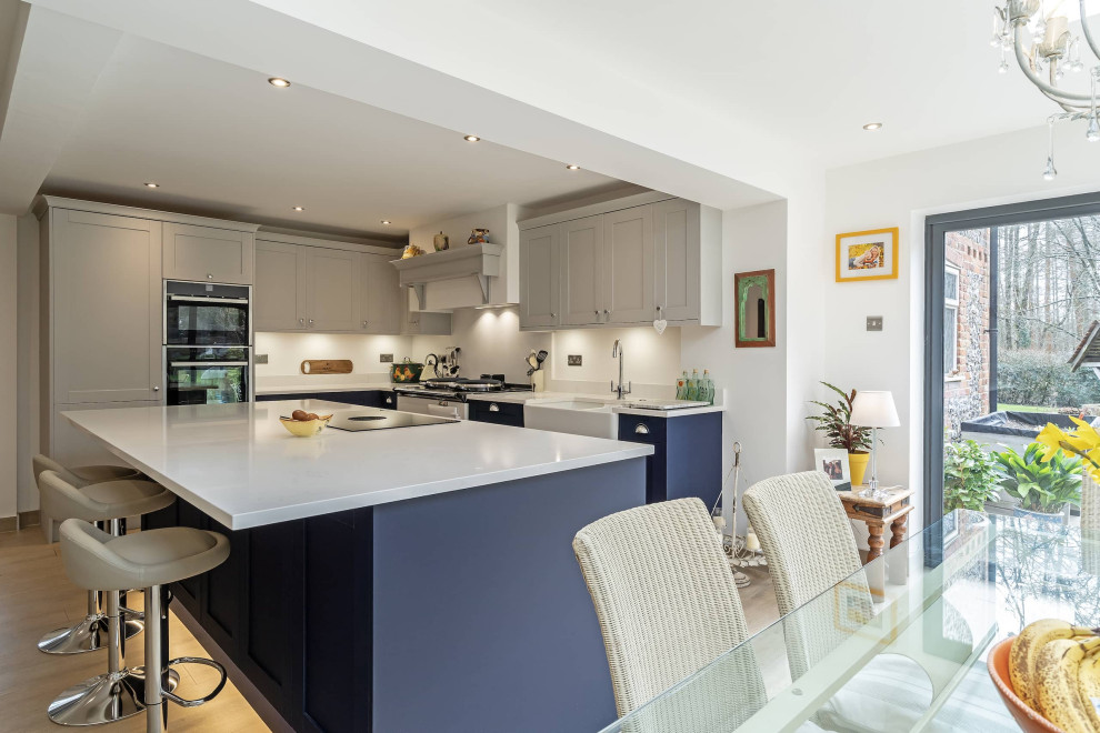 Photo of a mid-sized modern l-shaped eat-in kitchen in Oxfordshire with a farmhouse sink, shaker cabinets, white cabinets, quartzite benchtops, white splashback, black appliances, with island and white benchtop.