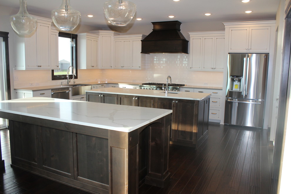 Example of a large transitional l-shaped dark wood floor enclosed kitchen design with a farmhouse sink, shaker cabinets, white cabinets, quartz countertops, white backsplash, subway tile backsplash, stainless steel appliances and an island