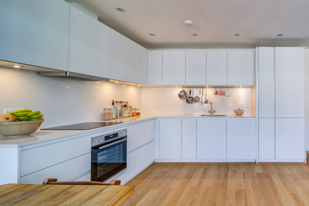 Photo of a mid-sized contemporary l-shaped eat-in kitchen in London with a double-bowl sink, flat-panel cabinets, white cabinets, quartzite benchtops, white splashback, black appliances, light hardwood floors, no island, brown floor and white benchtop.