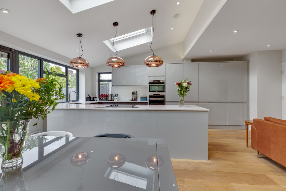 This is an example of a mid-sized contemporary u-shaped eat-in kitchen in London with an undermount sink, flat-panel cabinets, grey cabinets, quartz benchtops, white splashback, engineered quartz splashback, stainless steel appliances, medium hardwood floors, a peninsula, white benchtop and vaulted.