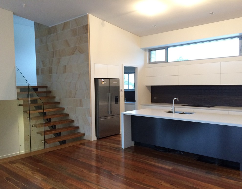 Contemporary galley open plan kitchen in Wollongong with an integrated sink, flat-panel cabinets, white cabinets, solid surface benchtops, blue splashback, glass sheet splashback, stainless steel appliances, medium hardwood floors, with island, brown floor and white benchtop.