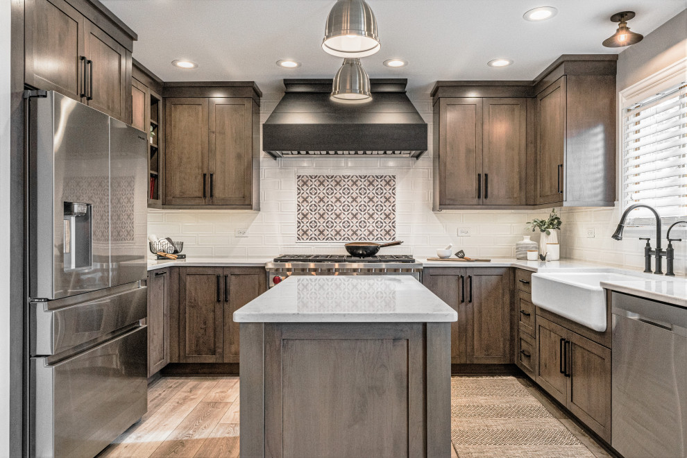 Mid-sized transitional u-shaped eat-in kitchen in Chicago with a farmhouse sink, shaker cabinets, brown cabinets, quartz benchtops, white splashback, porcelain splashback, stainless steel appliances, vinyl floors, with island, brown floor and white benchtop.