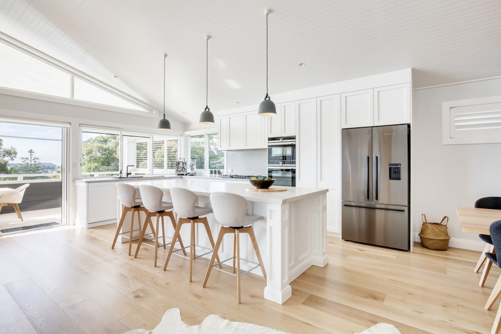 This is an example of a large beach style l-shaped open plan kitchen in Sydney with a drop-in sink, shaker cabinets, white cabinets, quartz benchtops, engineered quartz splashback, stainless steel appliances, light hardwood floors, with island, grey splashback, beige floor, white benchtop and vaulted.