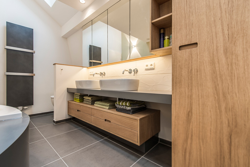 Photo of a small contemporary bathroom in Berlin with medium wood cabinets, solid surface benchtops, a corner tub, gray tile, ceramic tile, a vessel sink, a curbless shower, a wall-mount toilet, white walls and vinyl floors.