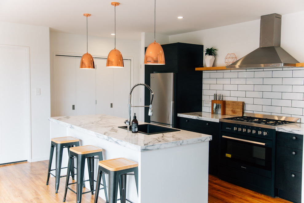 Contemporary galley kitchen in Christchurch with a drop-in sink, flat-panel cabinets, marble benchtops, white splashback, subway tile splashback, black appliances, medium hardwood floors, with island and orange floor.