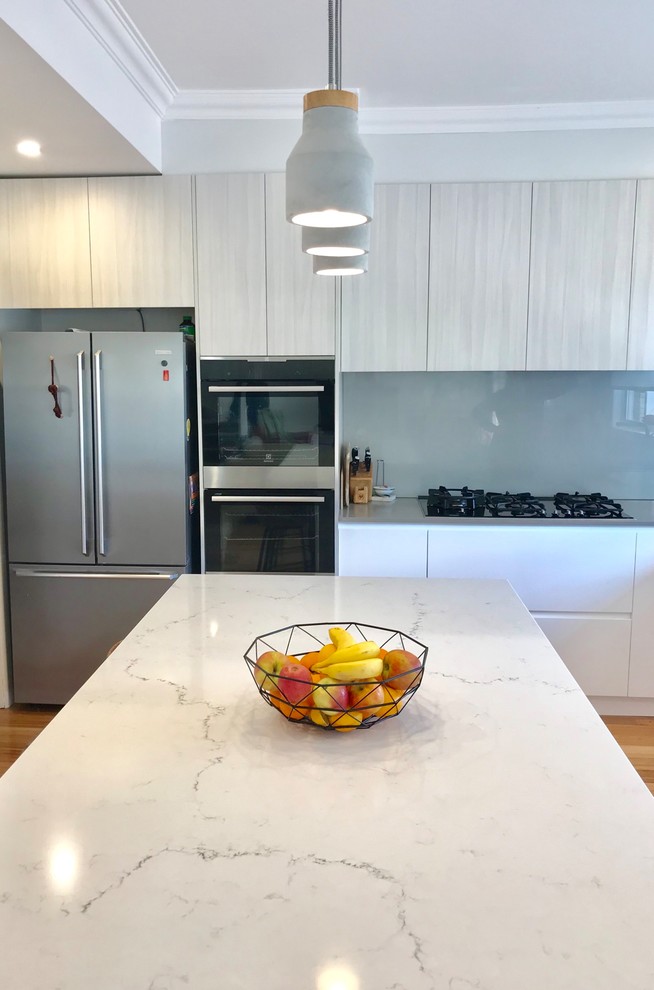 Design ideas for a mid-sized contemporary l-shaped eat-in kitchen in Melbourne with a double-bowl sink, white cabinets, marble benchtops, metallic splashback, glass sheet splashback, black appliances, light hardwood floors, with island, brown floor and grey benchtop.