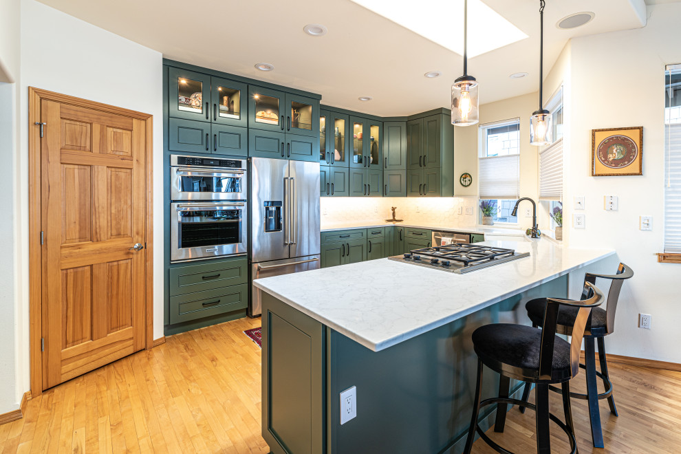 Example of a mid-sized transitional u-shaped medium tone wood floor and brown floor open concept kitchen design in Seattle with a farmhouse sink, green cabinets, white backsplash, stainless steel appliances, white countertops, flat-panel cabinets, quartz countertops, ceramic backsplash and a peninsula