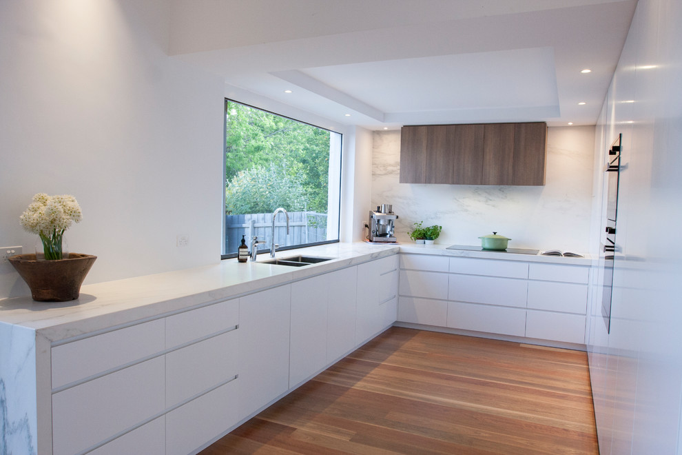 Mid-sized contemporary l-shaped kitchen in Melbourne with a double-bowl sink, flat-panel cabinets, white cabinets, marble benchtops, white splashback, marble splashback, stainless steel appliances, medium hardwood floors, multiple islands, brown floor and white benchtop.
