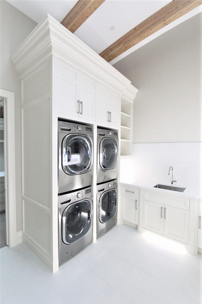 Large traditional single-wall dedicated laundry room in Calgary with an undermount sink, shaker cabinets, white cabinets, quartzite benchtops, white splashback, white walls, porcelain floors, a stacked washer and dryer, white floor, white benchtop and exposed beam.