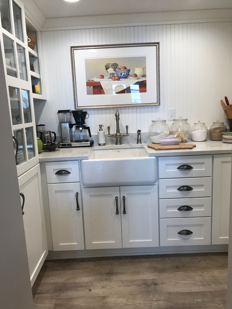 Photo of a large beach style l-shaped kitchen pantry in Other with a farmhouse sink, flat-panel cabinets, white cabinets, quartz benchtops, white splashback, timber splashback, stainless steel appliances, light hardwood floors, with island, grey floor and white benchtop.