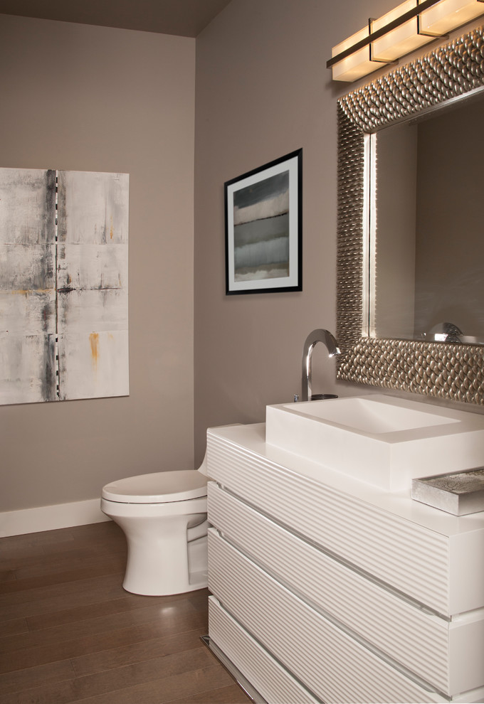 This is an example of a mid-sized contemporary powder room in Seattle with furniture-like cabinets, white cabinets, a two-piece toilet, white tile, grey walls, medium hardwood floors, a vessel sink, solid surface benchtops and grey floor.