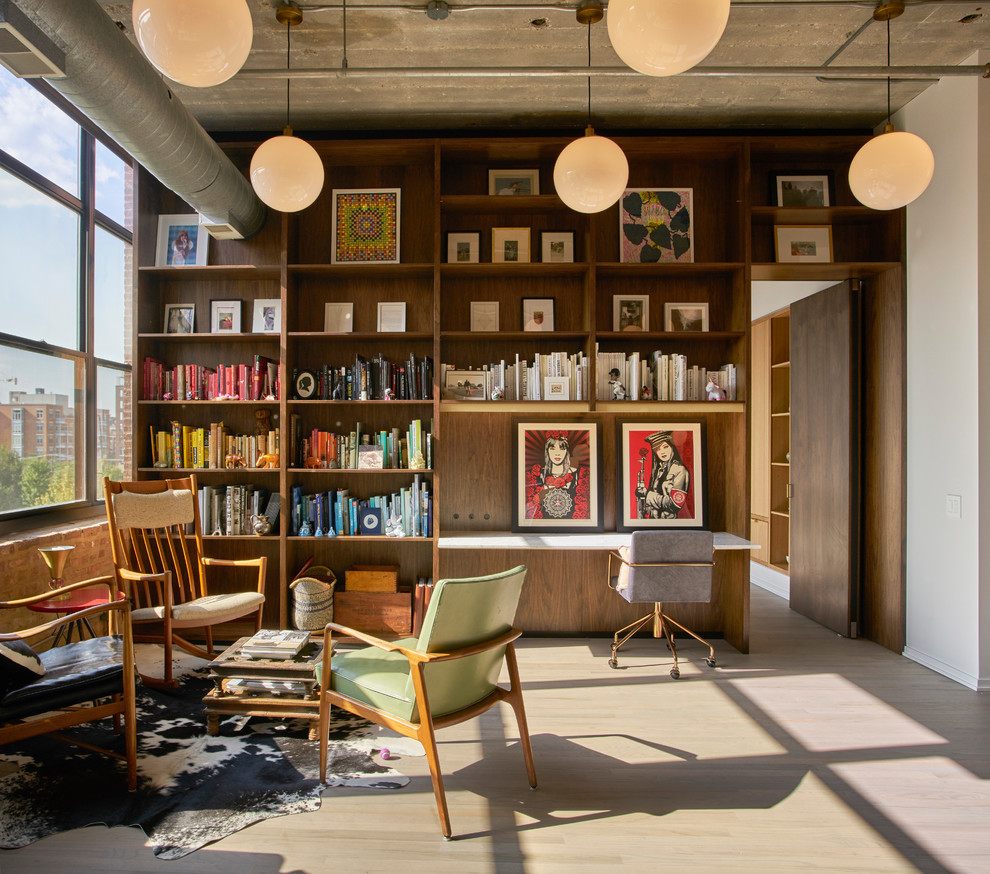 This is an example of an industrial home office in Chicago with a library, white walls, light hardwood floors, a built-in desk and grey floor.