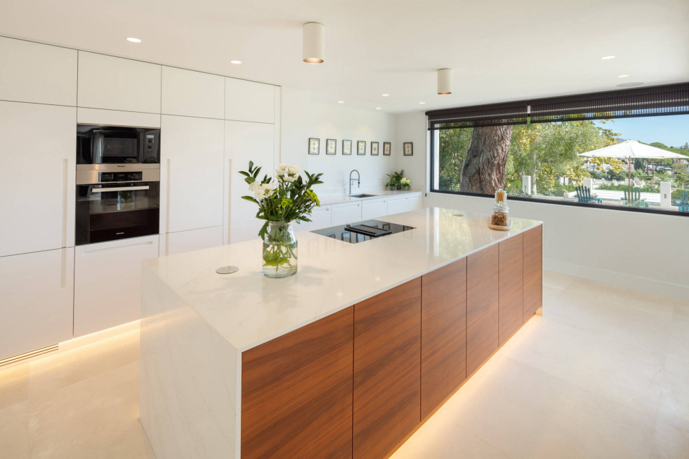 Large contemporary single-wall open plan kitchen in Malaga with a drop-in sink, flat-panel cabinets, light wood cabinets, black appliances, marble floors, with island and beige floor.