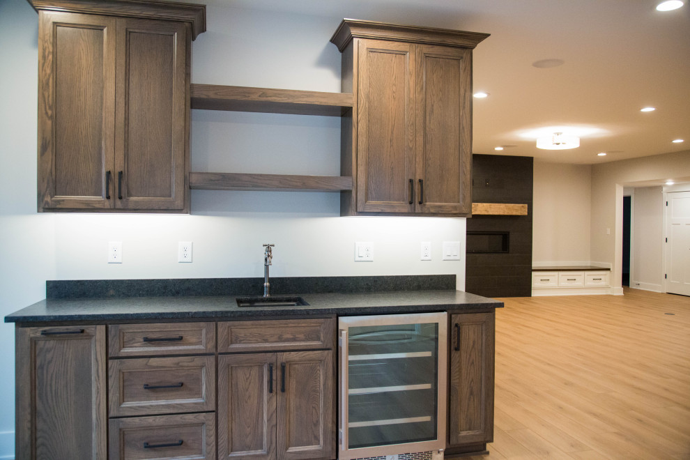 This is an example of a large traditional single-wall wet bar in Indianapolis with an undermount sink, recessed-panel cabinets, brown cabinets, granite benchtops, laminate floors, brown floor and black benchtop.