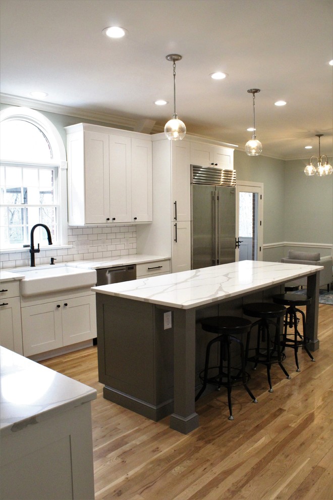 Example of a large transitional galley light wood floor eat-in kitchen design with a farmhouse sink, shaker cabinets, white cabinets, quartz countertops, white backsplash, ceramic backsplash, stainless steel appliances, an island and white countertops