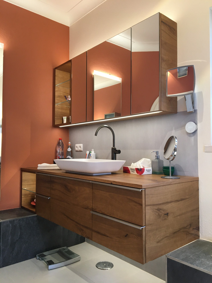 Photo of a large master bathroom in Nuremberg with dark wood cabinets, an open shower, a bidet, gray tile, ceramic tile, red walls, ceramic floors, a vessel sink, wood benchtops, grey floor, an open shower, brown benchtops, a single vanity and a floating vanity.