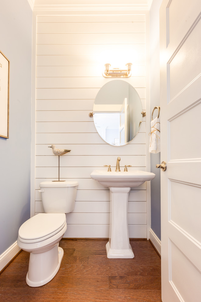 Photo of a mid-sized beach style powder room in Other with a two-piece toilet, white walls, medium hardwood floors, a pedestal sink and brown floor.