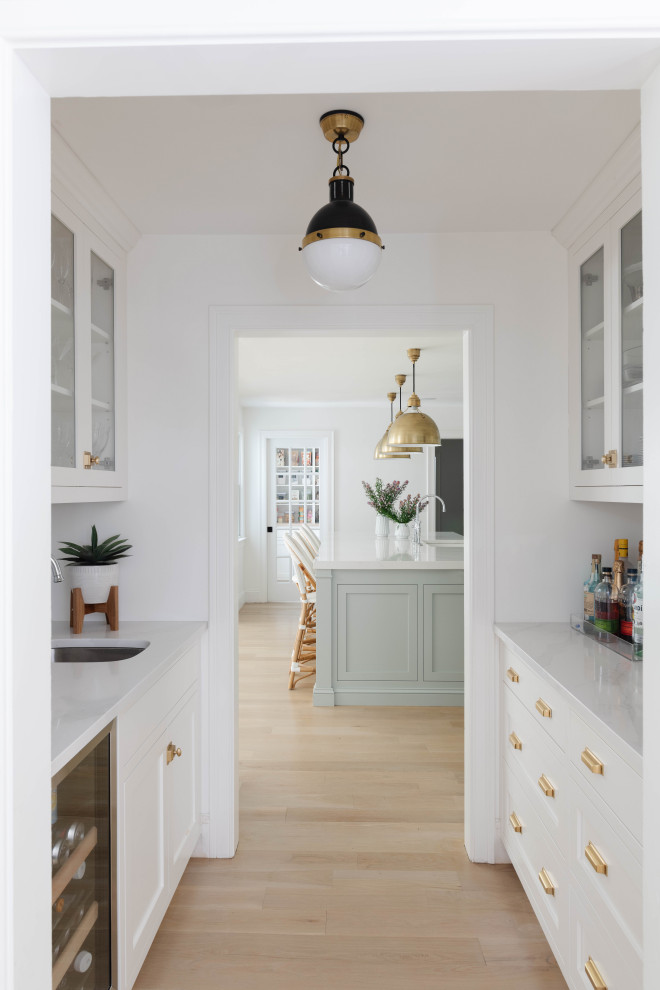 This is an example of a large classic galley kitchen pantry in Boston with a submerged sink, recessed-panel cabinets, white cabinets, engineered stone countertops, white splashback, ceramic splashback, stainless steel appliances, light hardwood flooring, an island and white worktops.