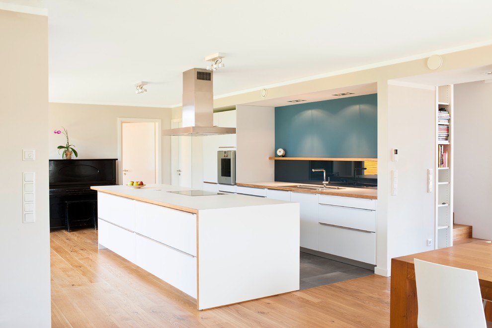 This is an example of a mid-sized contemporary single-wall open plan kitchen in Frankfurt with an undermount sink, flat-panel cabinets, white cabinets, wood benchtops, blue splashback, glass sheet splashback, panelled appliances, medium hardwood floors and with island.