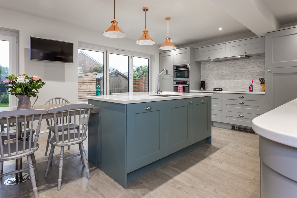 Photo of a medium sized eclectic l-shaped kitchen/diner in Hampshire with a submerged sink, shaker cabinets, grey cabinets, composite countertops, grey splashback, stainless steel appliances, laminate floors, an island, beige floors and white worktops.