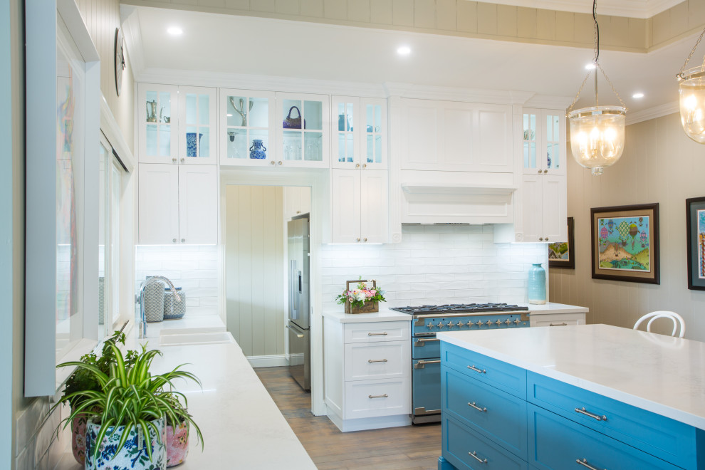 This is an example of an expansive classic l-shaped kitchen pantry in Brisbane with a belfast sink, shaker cabinets, white cabinets, engineered stone countertops, white splashback, metro tiled splashback, coloured appliances, medium hardwood flooring, an island and white worktops.