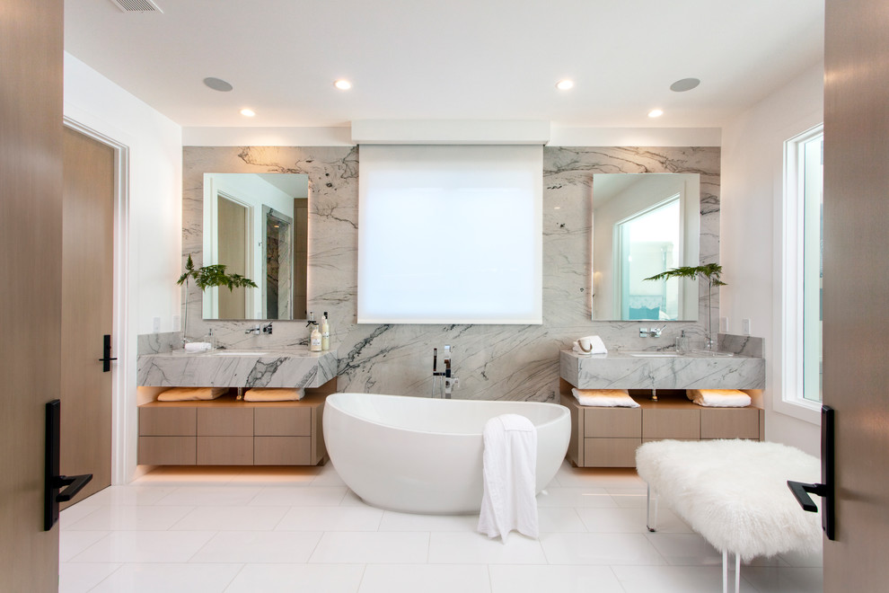 Photo of a large beach style master bathroom in Orange County with flat-panel cabinets, brown cabinets, a freestanding tub, gray tile, marble, white walls, porcelain floors, an undermount sink, marble benchtops, white floor and grey benchtops.