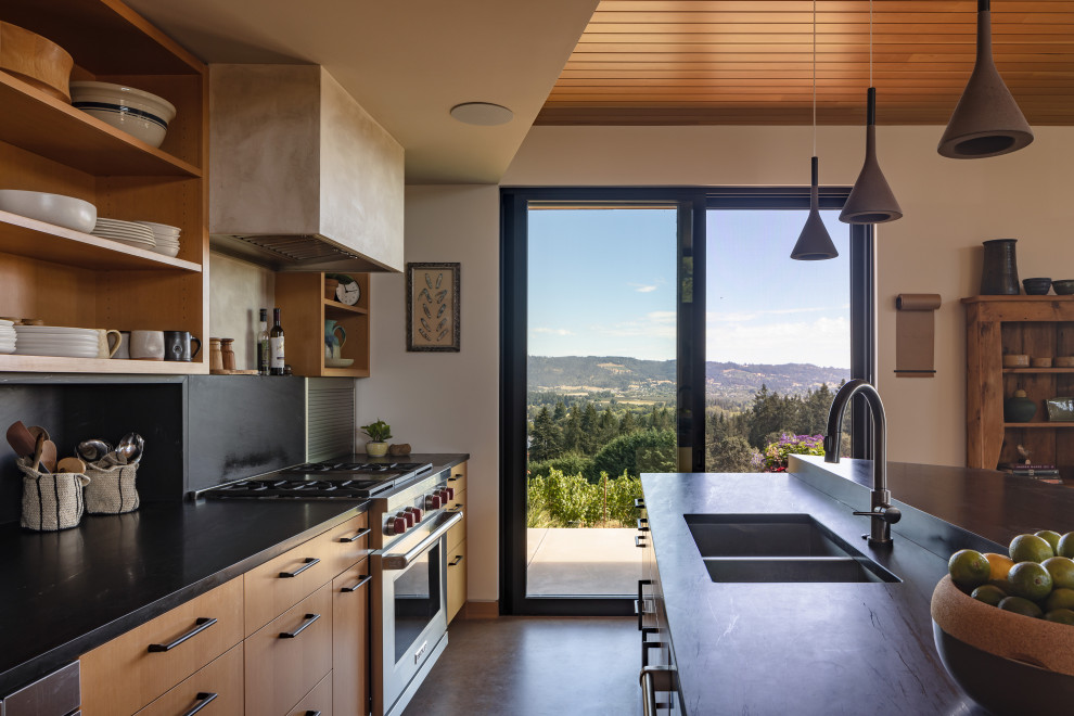 This is an example of a mid-sized modern single-wall eat-in kitchen in Portland with light wood cabinets, soapstone benchtops, black splashback, stainless steel appliances, concrete floors, with island, black benchtop, wood, an undermount sink, flat-panel cabinets, stone slab splashback and grey floor.