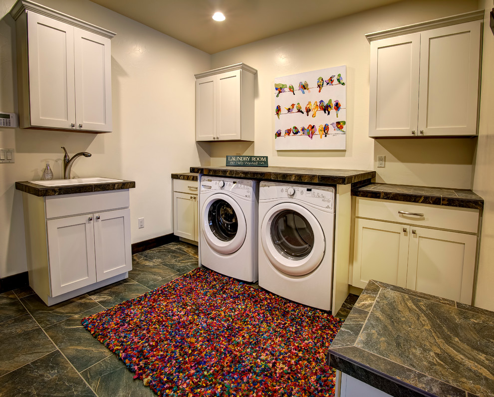 This is an example of a large transitional u-shaped dedicated laundry room in Other with a drop-in sink, shaker cabinets, white cabinets, tile benchtops, white walls, slate floors and a side-by-side washer and dryer.