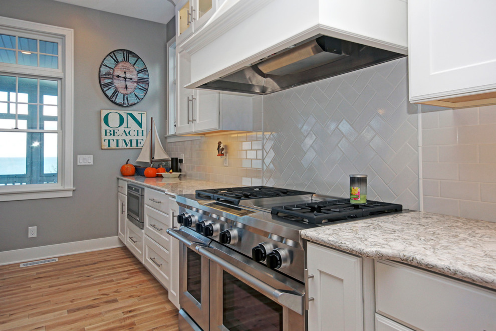 Photo of a beach style open plan kitchen in Grand Rapids with shaker cabinets, white cabinets, grey splashback, subway tile splashback, stainless steel appliances and light hardwood floors.