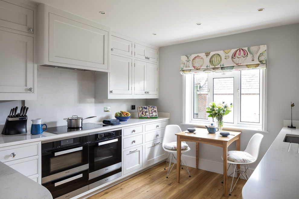 Design ideas for a transitional u-shaped eat-in kitchen in Other with an undermount sink, beaded inset cabinets, white cabinets, white splashback, stone slab splashback, black appliances, medium hardwood floors, no island, brown floor and white benchtop.