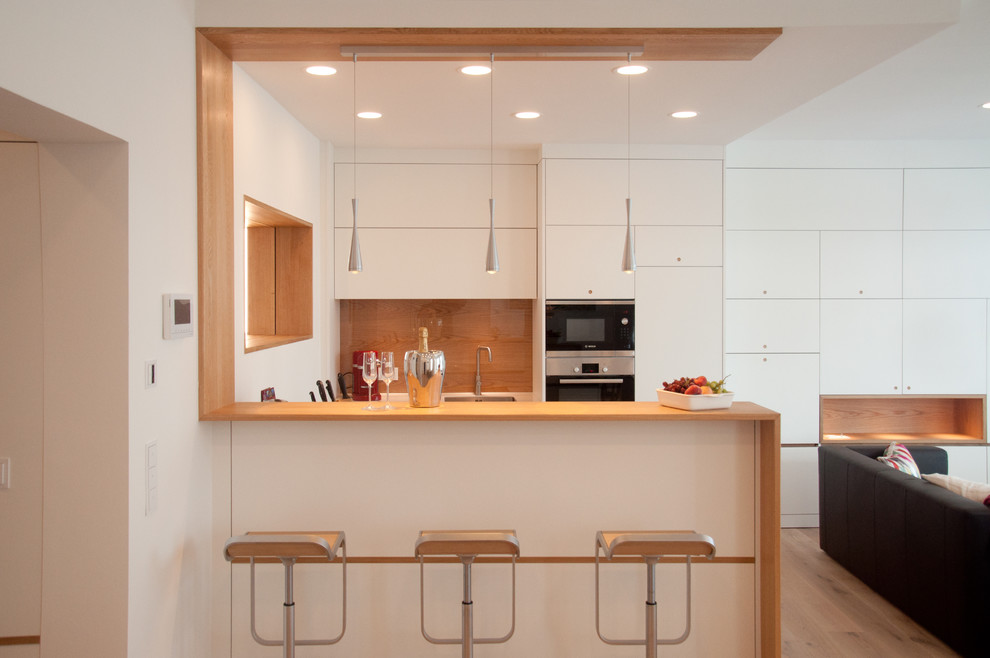 This is an example of a small contemporary u-shaped open plan kitchen in Hamburg with an undermount sink, flat-panel cabinets, white cabinets, light hardwood floors, a peninsula and brown splashback.