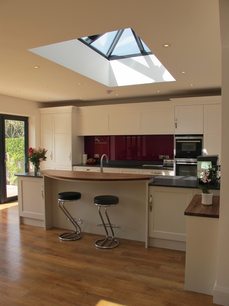 Photo of a mid-sized contemporary l-shaped open plan kitchen in Surrey with a drop-in sink, flat-panel cabinets, granite benchtops, red splashback, glass tile splashback, stainless steel appliances, medium hardwood floors and with island.