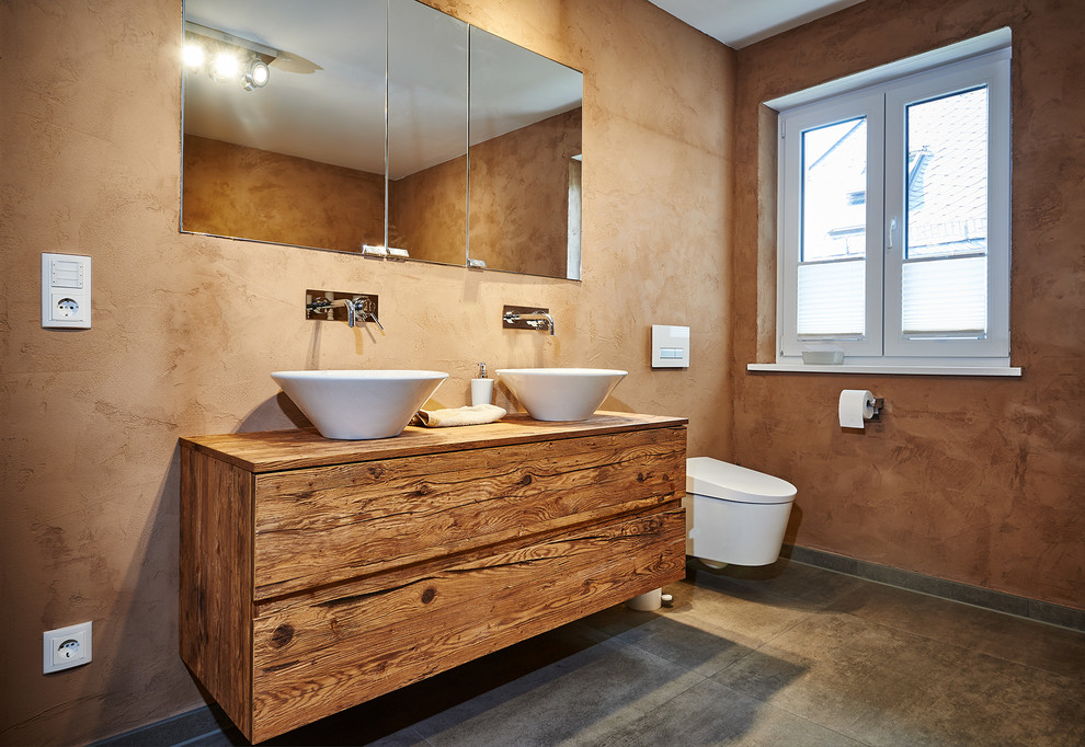 Inspiration for a contemporary bathroom in Munich with a vessel sink, medium wood cabinets, wood benchtops, brown walls, flat-panel cabinets, a wall-mount toilet, gray tile and stone slab.