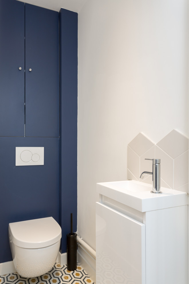 Photo of a small contemporary powder room in Paris with beaded inset cabinets, blue cabinets, a wall-mount toilet, white tile, ceramic tile, blue walls, cement tiles, a wall-mount sink, yellow floor and white benchtops.