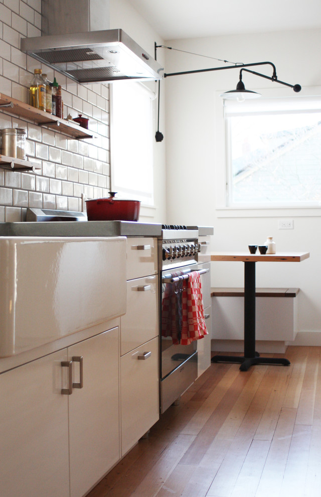 This is an example of a mid-sized industrial galley eat-in kitchen in Portland with a farmhouse sink, flat-panel cabinets, white cabinets, concrete benchtops, white splashback, subway tile splashback, stainless steel appliances, light hardwood floors and no island.