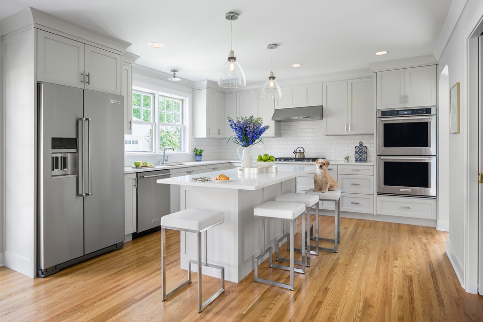 Photo of a mid-sized transitional l-shaped open plan kitchen in Minneapolis with an undermount sink, shaker cabinets, white cabinets, white splashback, subway tile splashback, stainless steel appliances, light hardwood floors, with island and brown floor.