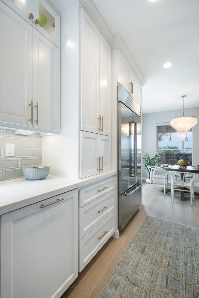 This is an example of a beach style kitchen in Houston with a farmhouse sink, white cabinets, quartz benchtops, blue splashback, ceramic splashback, stainless steel appliances, light hardwood floors, with island and white benchtop.