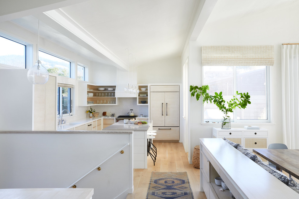 Beach style l-shaped kitchen in San Francisco with an undermount sink, flat-panel cabinets, light wood cabinets, white splashback, panelled appliances, light hardwood floors, with island, beige floor and white benchtop.