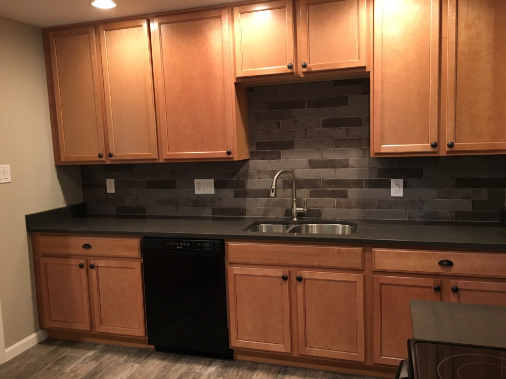 Mid-sized traditional l-shaped kitchen pantry in Baltimore with a double-bowl sink, shaker cabinets, medium wood cabinets, solid surface benchtops, grey splashback, subway tile splashback, black appliances, vinyl floors, no island, grey floor and black benchtop.