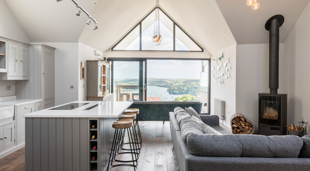 This is an example of a mid-sized beach style open plan kitchen in Devon with a farmhouse sink, shaker cabinets, grey cabinets, solid surface benchtops, light hardwood floors, with island and brown floor.