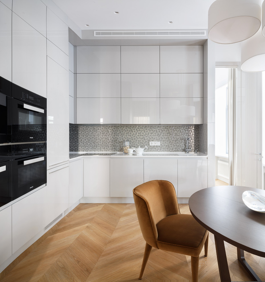 Mid-sized contemporary l-shaped eat-in kitchen in Moscow with an undermount sink, flat-panel cabinets, white cabinets, black appliances, light hardwood floors, quartzite benchtops, metallic splashback, mosaic tile splashback and no island.