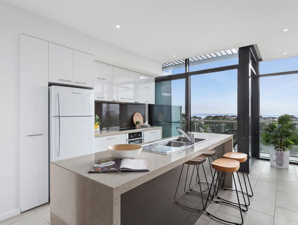 This is an example of a contemporary galley kitchen in Geelong with a double-bowl sink, flat-panel cabinets, white cabinets, black splashback, glass sheet splashback, stainless steel appliances, with island, beige floor and beige benchtop.