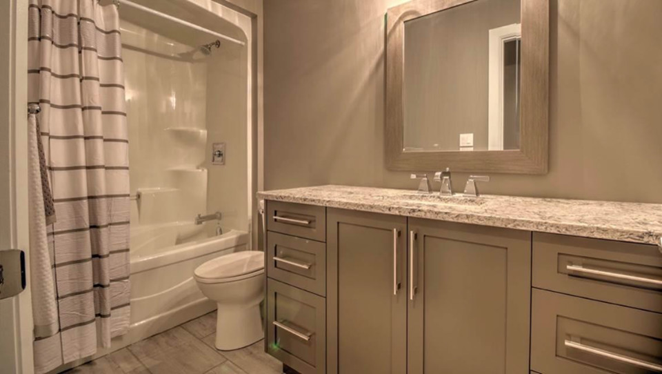 Example of a small transitional 3/4 medium tone wood floor and brown floor alcove bathtub design with shaker cabinets, gray cabinets, a one-piece toilet, brown walls, an undermount sink, granite countertops and gray countertops